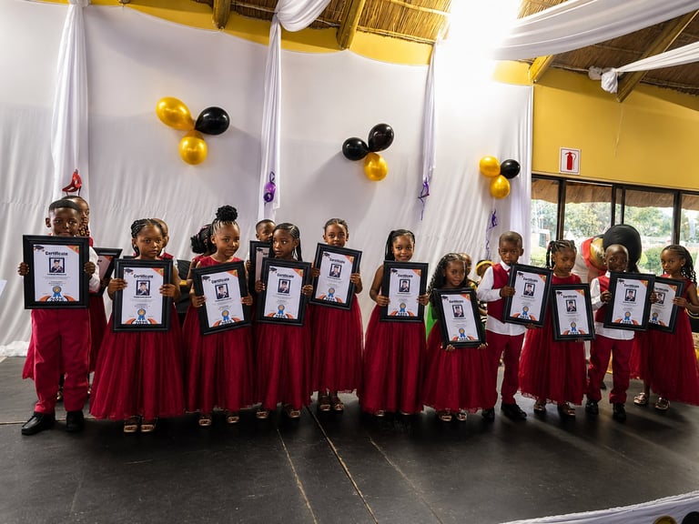 Group of children in red skirts holding framed certificates with gold and black balloons hanging above them on stage