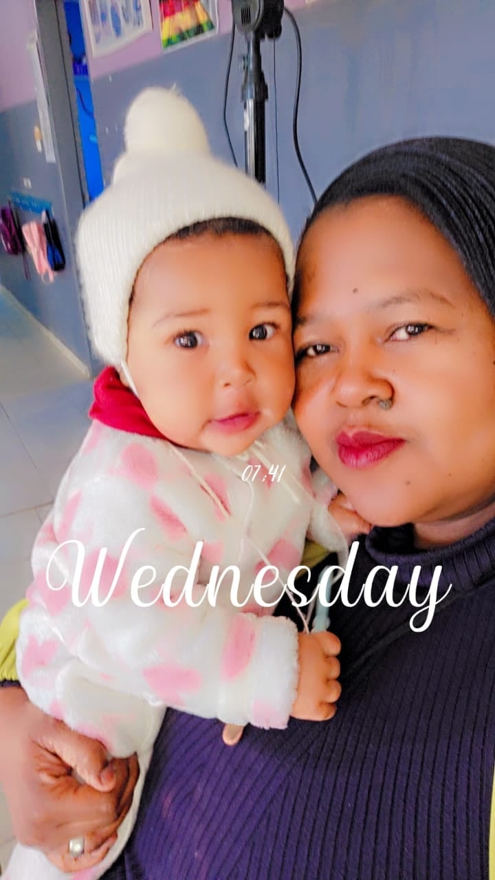 Woman holding a baby wearing a white beanie and pink outfit, both looking at camera indoors