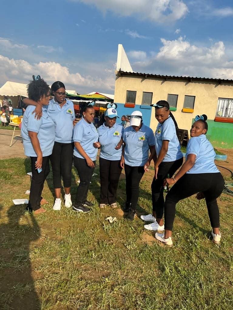 A group of people wearing matching light blue shirts and caps huddle together on a grassy field with colorful buildings in the background under a blue sky