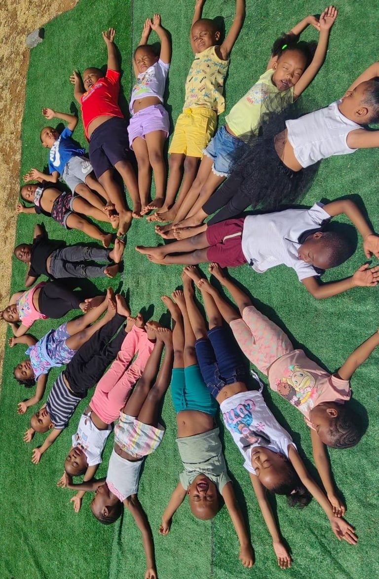 Group of children lying in a circle on green turf with hands raised toward center, viewed from above
