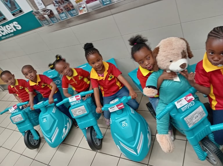 Group of children in red and yellow uniforms posing with toy turquoise scooters and a plush teddy bear mascot indoors