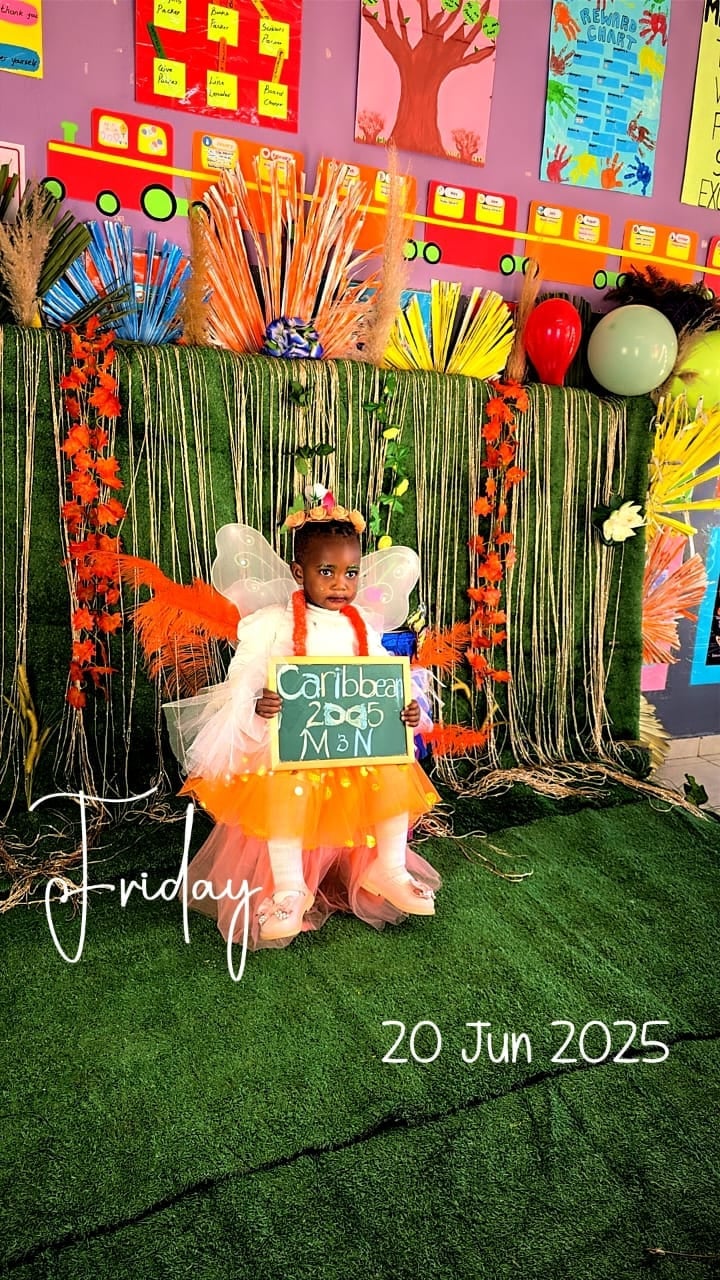 Young child in fairy costume with wings holding Caribbean Man sign at classroom birthday display, dated Friday 20 June 2025