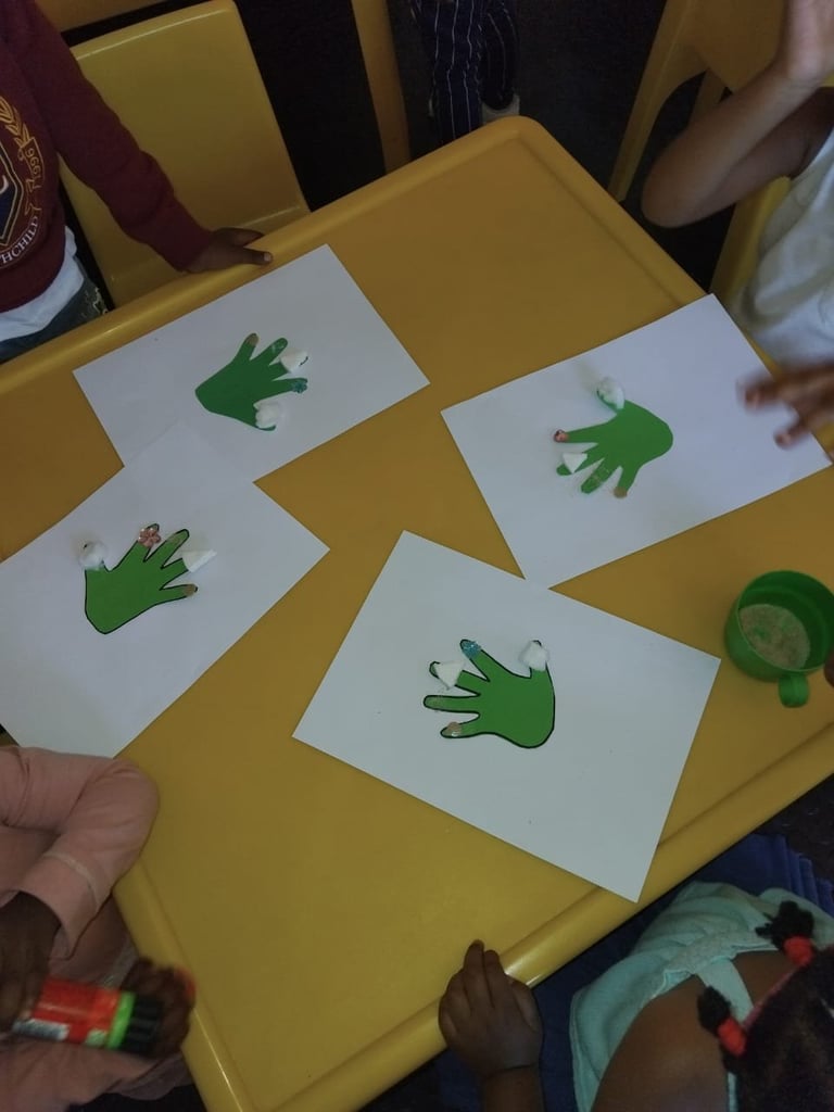 Green handprint art cards arranged on a yellow table with childrens hands visible around them during a craft activity