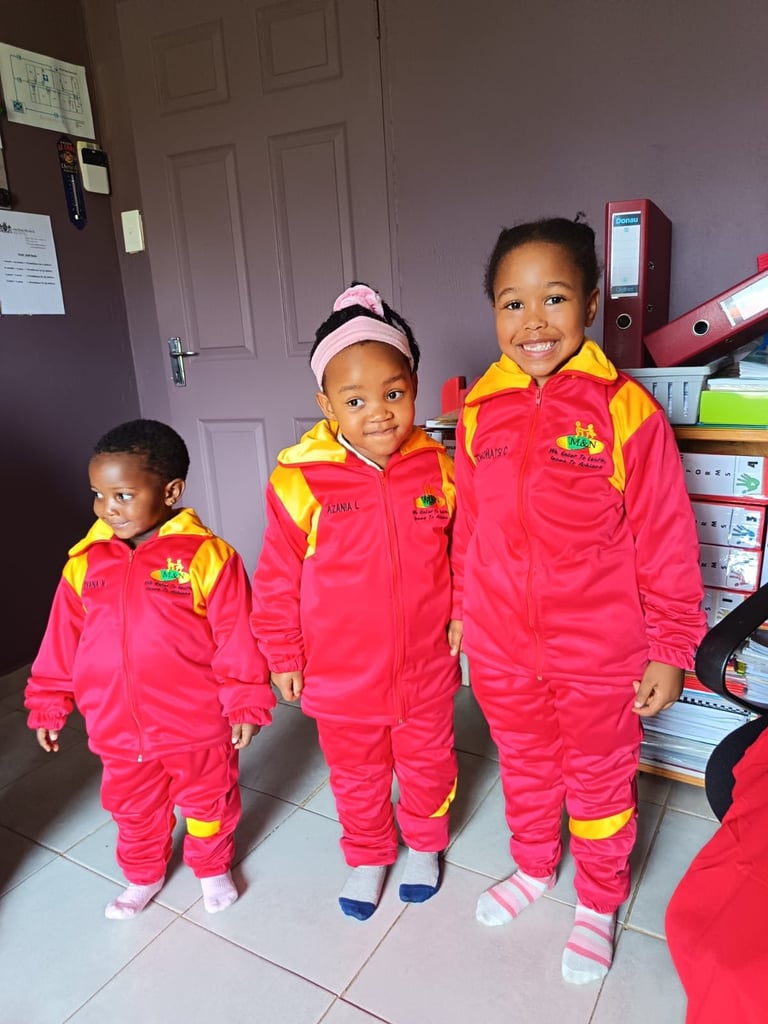 Three young children wearing matching red and yellow jumpsuits with logos stand together indoors in a classroom setting