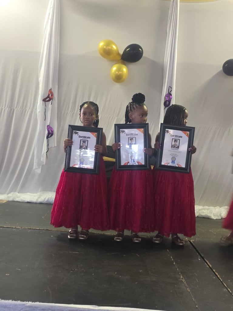 Three young girls in red skirts holding framed certificates on stage with white backdrop and yellow balloons
