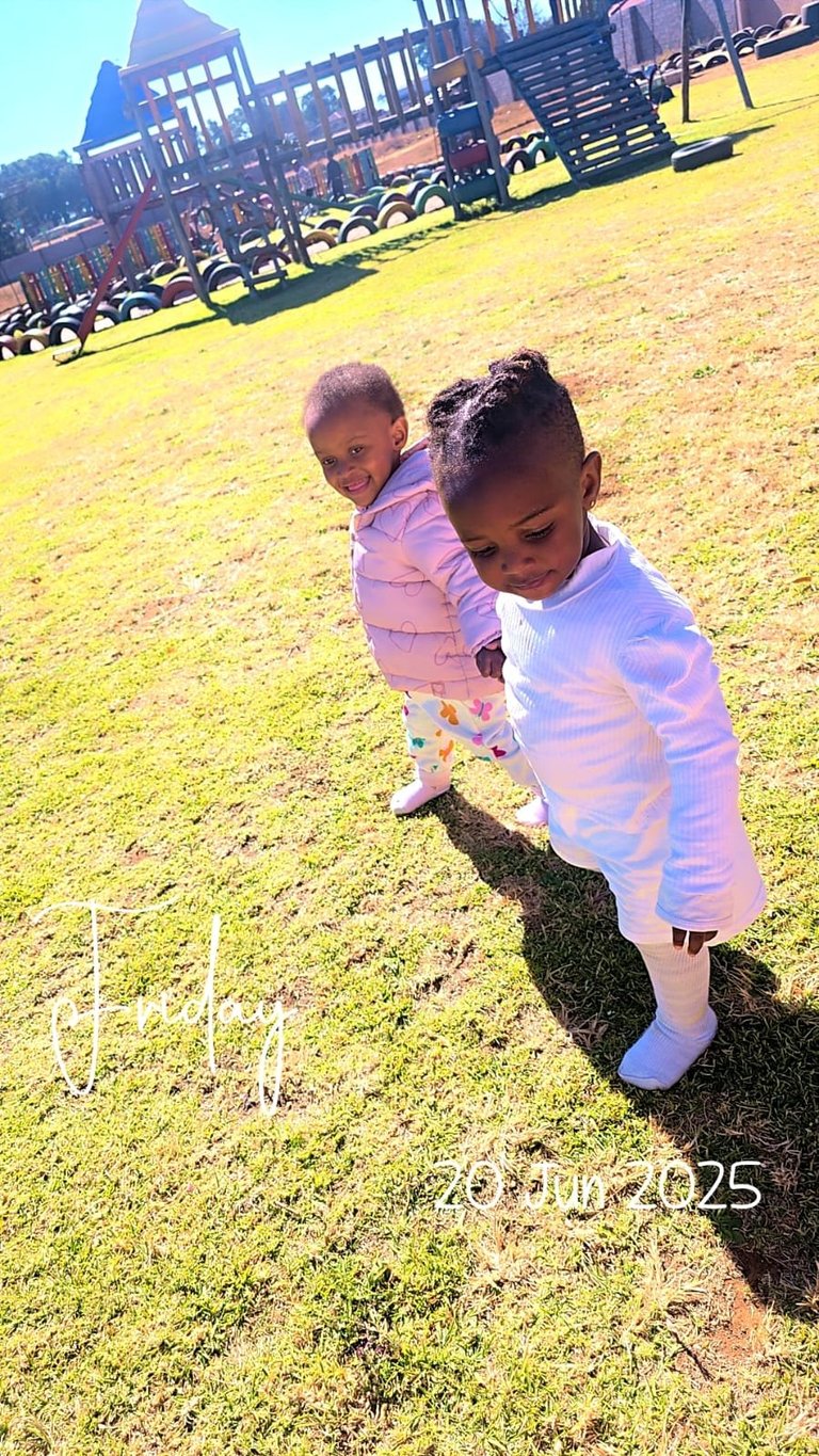Two young children playing on a sunny grass field at a farm with wooden structures and tractors in the background