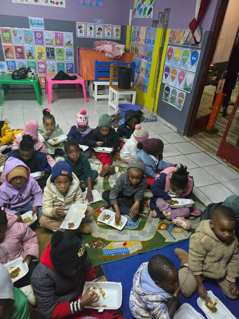 Group of children sitting on the floor eating lunch in a colorful classroom with artwork displayed on walls
