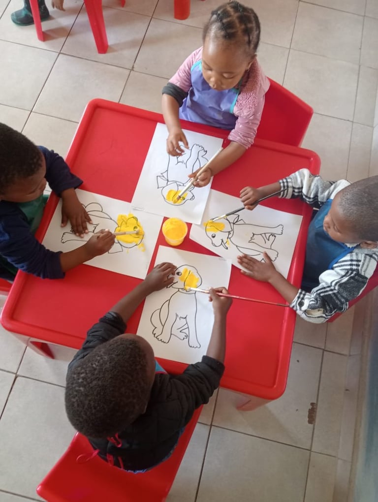 Young children coloring drawings together at a red table in a classroom or daycare setting